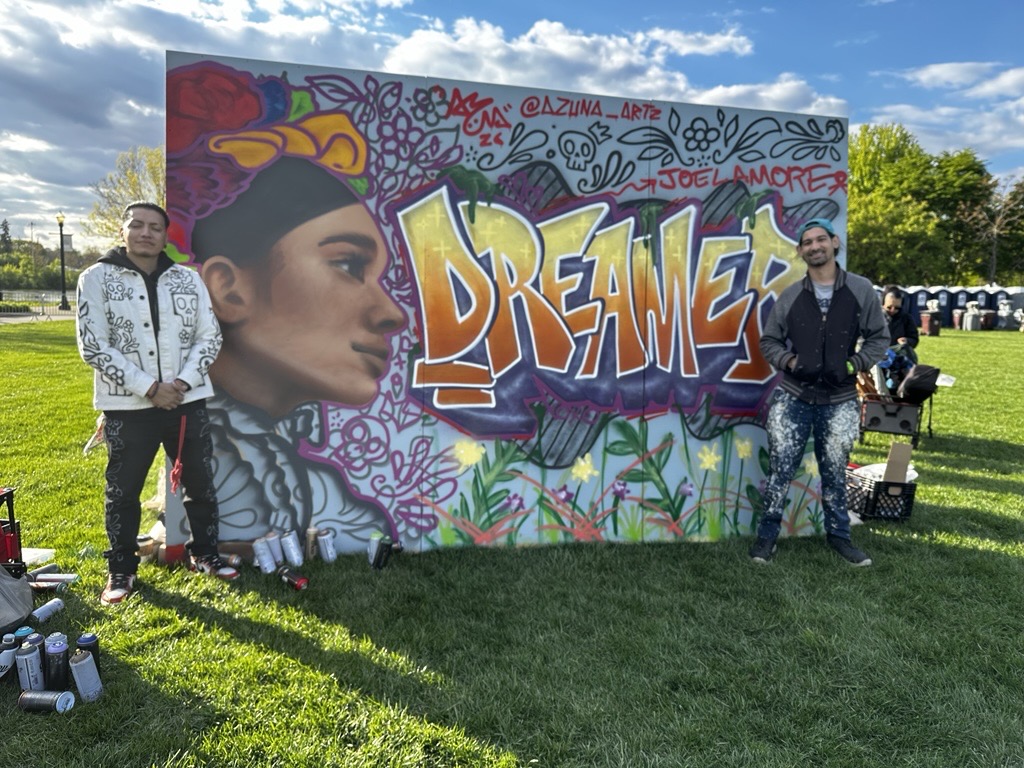 Two artists stand beside a large mural during Viva La Fest at Festival Park in downtown Elgin. The colorful artwork features a portrait, floral designs, and the word “DREAMER” in bold graffiti-style lettering, showcasing live community art and cultural expression.
