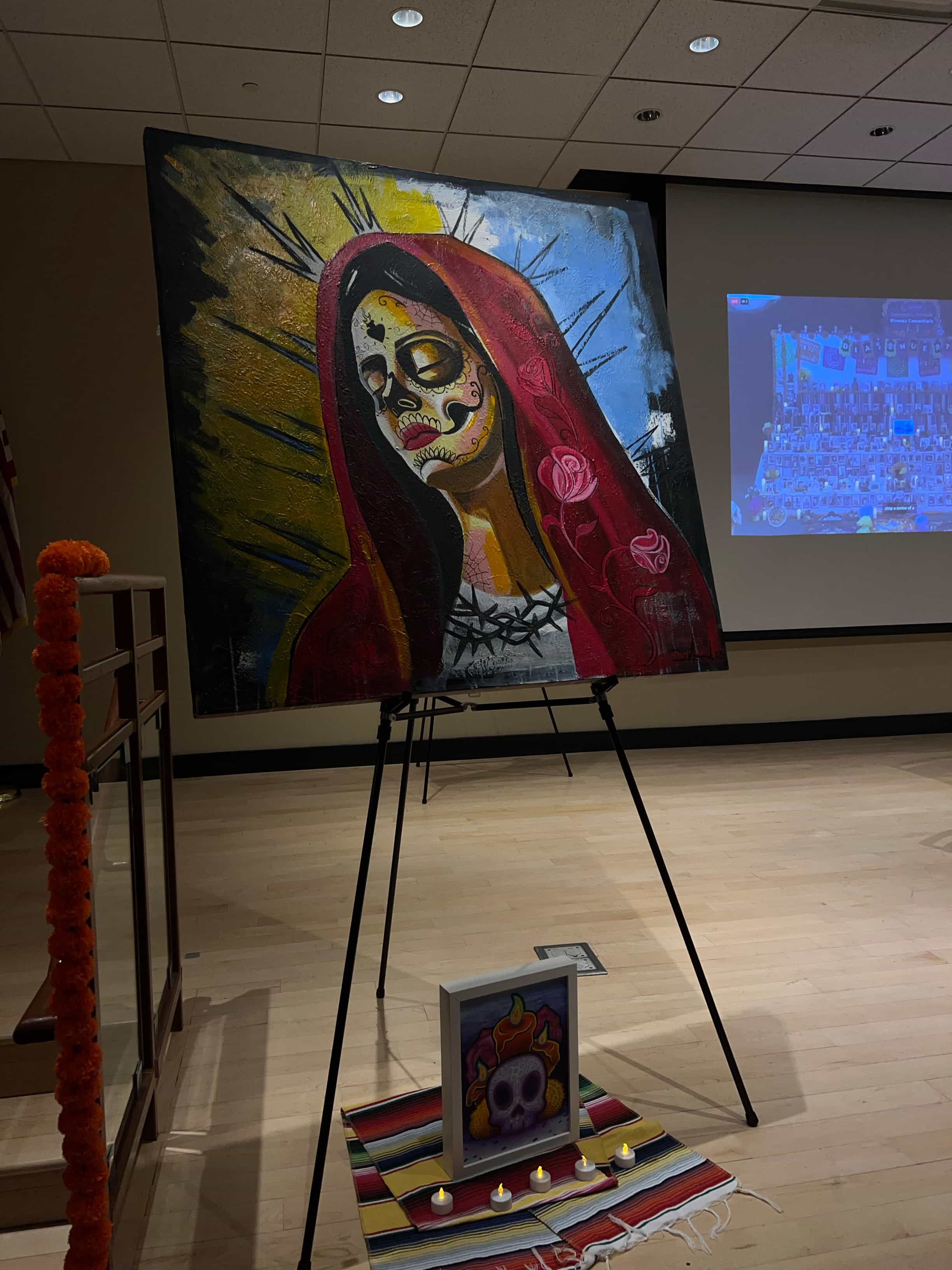 Large-scale art work painting inspired by Día de los Muertos traditions, displayed on an easel in the library. The artwork depicts a figure with a skull face and red mantle, accompanied by a small altar featuring candles and a framed artistic illustration at the base.
