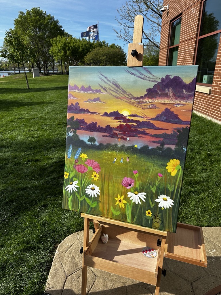 Artwork painting displayed outdoors on an easel, depicting a field of wildflowers beneath a colorful sunset sky. The artwork features vibrant shades of purple, orange, and yellow, with flowers in the foreground and a natural landscape in the background.